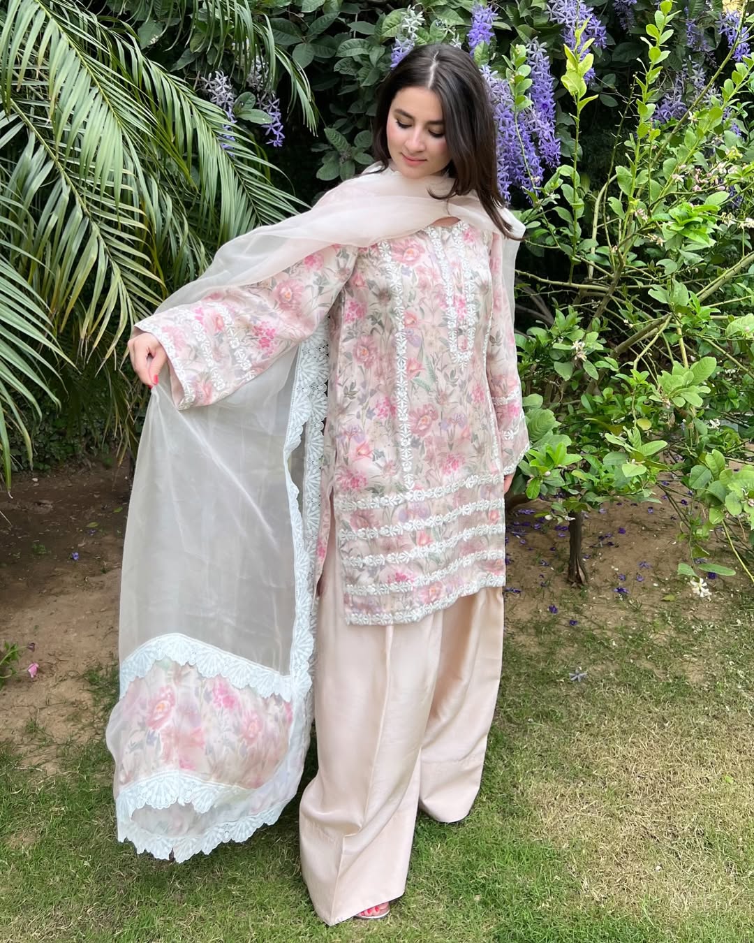 A woman standing outdoors wearing a traditional three-piece organza fabric outfit with floral embroidery and a dupatta.