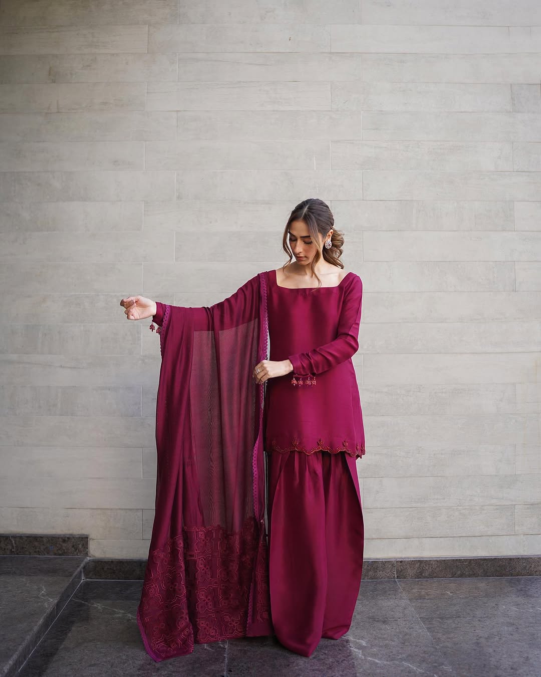 A woman wearing a burgundy long farshi shalwar set with handcrafted details on the ghera and sleeves, standing against a grey background.
