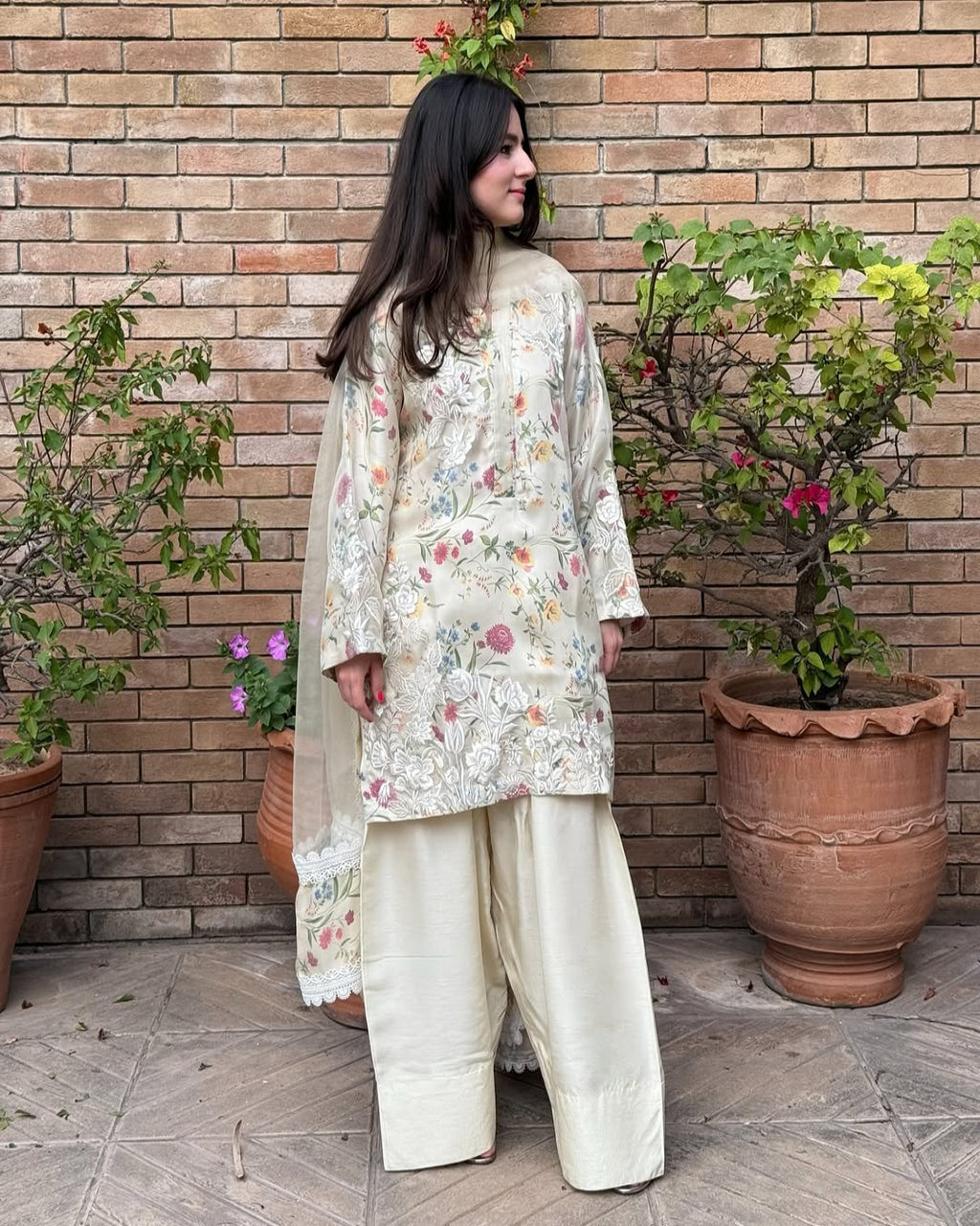 A woman standing against a brick wall with potted plants to her sides, wearing a traditional Pakistani Gulzar with floral embroidery and organza fabric.